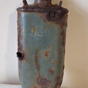 May include: A vintage, teal metal flask, heavily rusted and showing signs of age. The flask has a rounded base and a narrow neck. The surface is marked with holes and the text "H.G.S. BRUN 1940". The flask is likely from the 1940s.