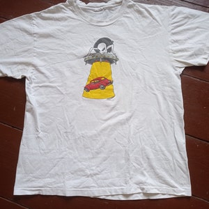 Könnte beinhalten: Weißes T-Shirt mit einem Aufdruck einer fliegenden Untertasse, die ein rotes Auto entführt. Die Untertasse hat einen schwarzen Außerirdischen im Inneren und das Auto befindet sich auf einem gelben Strahl.
