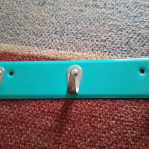 Peut inclure: Un porte-manteau en métal turquoise avec trois crochets. Les crochets sont argentés et ont une forme arrondie.