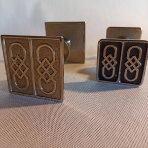 May include: Three silver metal cabinet knobs with a square shape and a Celtic knot design. The knobs have a silver finish and are in focus.