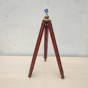 May include: A red wooden tripod with a metal top and a yellow base. The tripod is standing on a light brown surface.