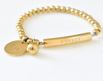 May include: Gold beaded bracelet with a cylindrical bar engraved with Roman numerals and a round charm with the name "Cory". The bracelet features a gold sphere charm and a gold disc charm. The bracelet is on a white background.