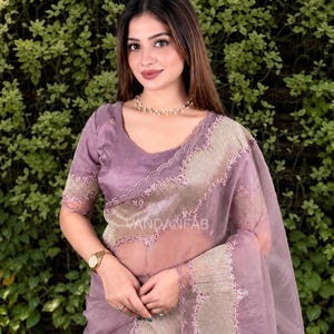 May include: A mauve organza saree featuring a scalloped border with intricate floral embroidery and a metallic gold panel. The garment includes a matching short-sleeved blouse with a V-neckline and the text VANDANFAB printed on the fabric.