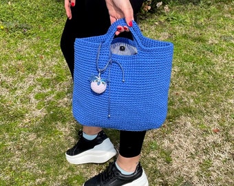 Könnte beinhalten: Eine blaue gehäkelte Tragetasche mit einem rosa-blauen Anhänger, der an der Henkel hängt. Die Tasche hat ein schwarz-weiß gestreiftes Futter.