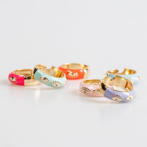 May include: Five gold hoop earrings with colourful enamel and eye designs. The earrings are in shades of pink, blue, green, and orange.
