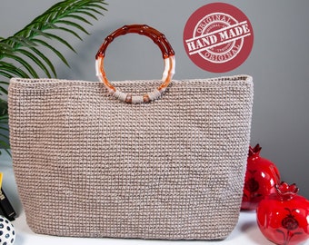 May include: A large, beige crocheted tote bag with a brown and white bamboo handle. The bag is sitting on a white table with a plant, books, and red ceramic pomegranates in the background. The bag has a red and white "ORIGINAL HAND MADE ORIGINAL" stamp on it.