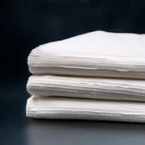 May include: A stack of three white cotton napkins with a textured pattern. The napkins are folded neatly and are arranged on a dark background.