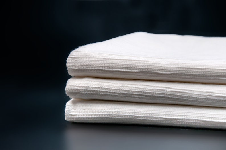 May include: A stack of three white cotton napkins with a textured pattern. The napkins are folded neatly and are arranged on a dark background.