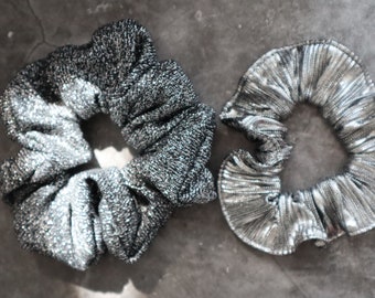Op de afbeelding: Twee glinsterende scrunchies in de afbeelding. Eén is donker zilver met een getextureerde uitstraling, de andere is licht zilver met een geplooid ontwerp. Beide zijn cirkelvormig en gemaakt van een glanzende stof, perfect om te accessoriseren.