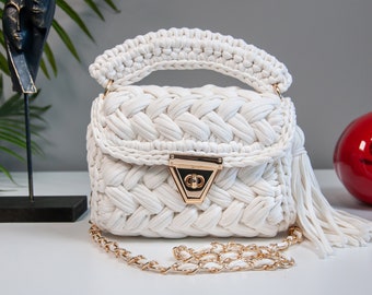 May include: A white crocheted handbag with a gold chain strap and a gold clasp. The bag has a tassel on the side.