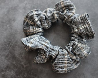 May include: A gray and beige plaid scrunchie. The hair accessory is made of a soft, textured fabric. The scrunchie is circular and gathered, with a classic plaid pattern. The background is a solid gray.