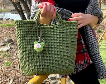May include: A green crocheted tote bag with a small crocheted bear charm. The bag has a simple design and is perfect for everyday use.