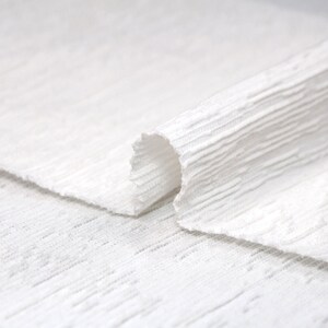 May include: White textured fabric folded over itself, showing the texture of the fabric.
