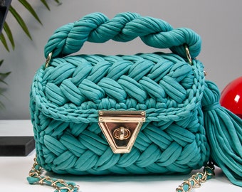 May include: A teal blue woven handbag with a gold chain strap and a gold clasp. The bag has a braided handle and a tassel.