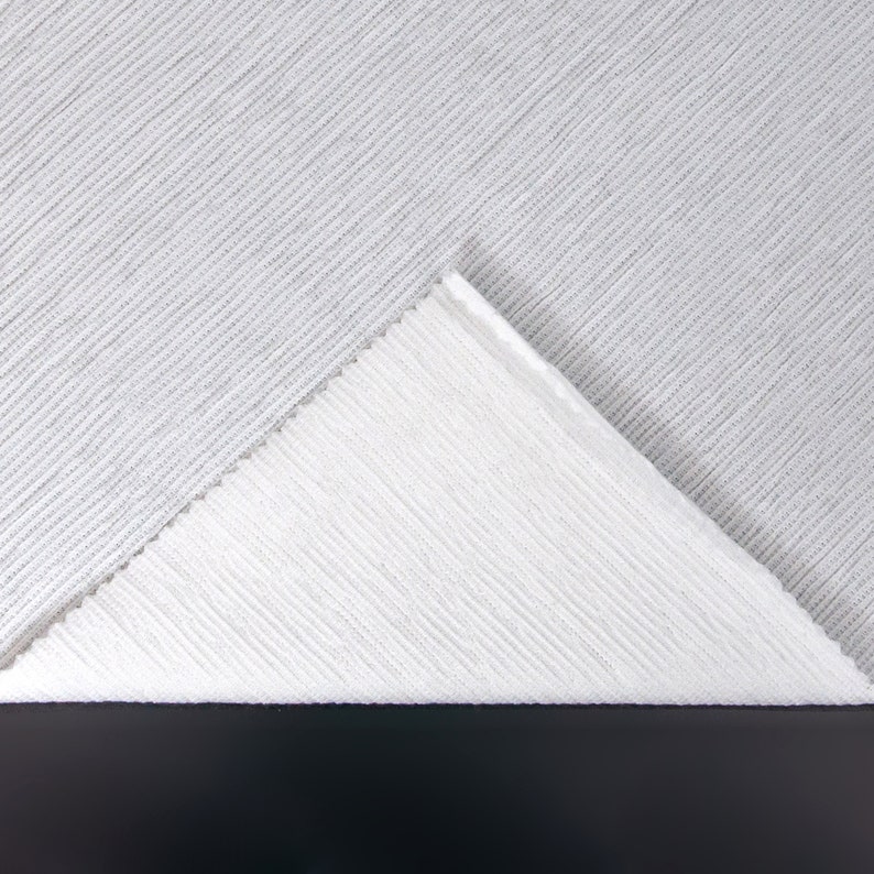 May include: Close-up of two white ribbed fabrics, one folded over the other, creating a triangular shape. The fabric has a subtle texture and a soft, light appearance.