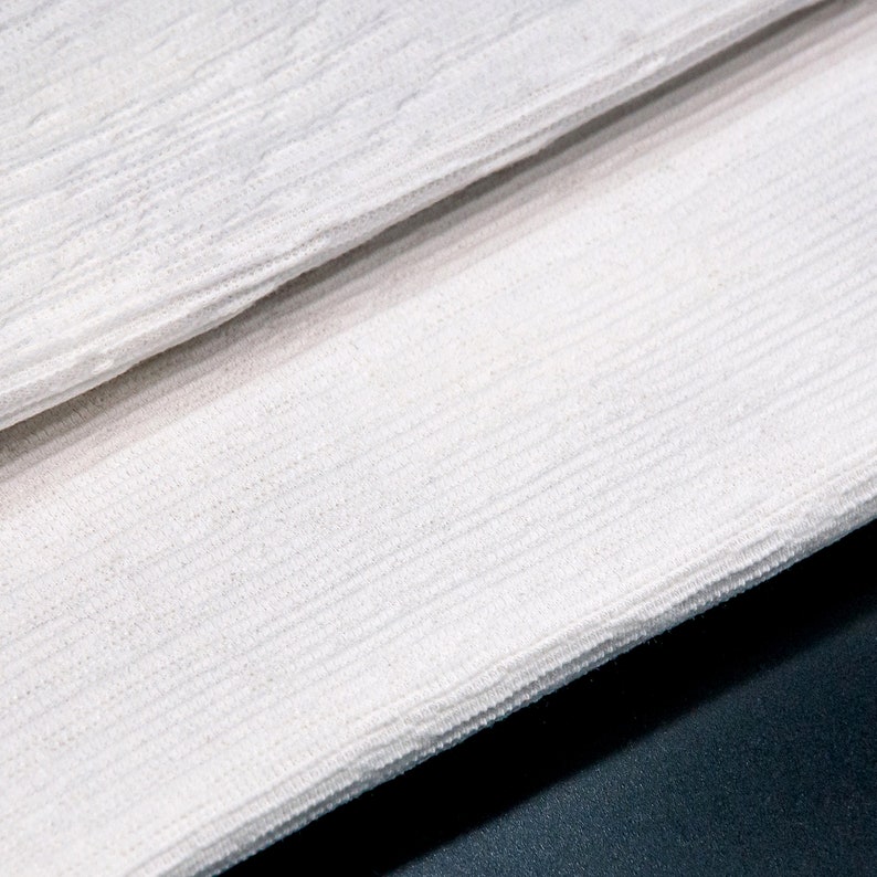 May include: Close-up of white fabric with a textured, woven pattern. The fabric is folded in half, creating a diagonal line.