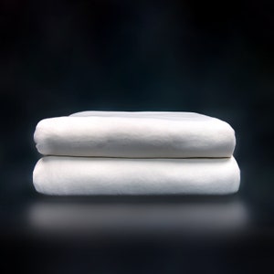 May include: Two white folded cotton sheets stacked on top of each other against a dark background.