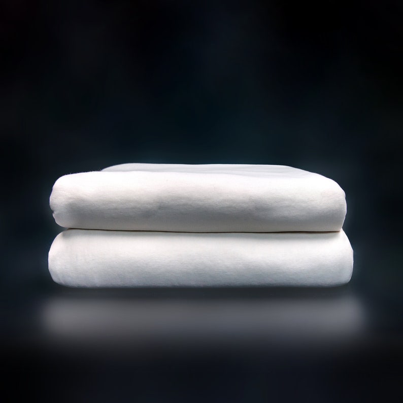May include: Two white folded cotton sheets stacked on top of each other against a dark background.