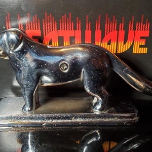 May include: A silver metal nutcracker shaped like a dog. The dog is standing on a base with a smooth, reflective surface. The dog's tail is curled up and the nutcracker has a screw on the side.