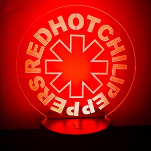 May include: A red illuminated sign featuring the words "Red Hot Chili Peppers" encircling a white asterisk symbol. The sign is circular and sits on a black base, with a red background.
