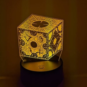 May include: A 3D printed lamp shaped like a cube with a complex geometric pattern. The lamp is lit up in a warm yellow color and has a black base with a power switch.