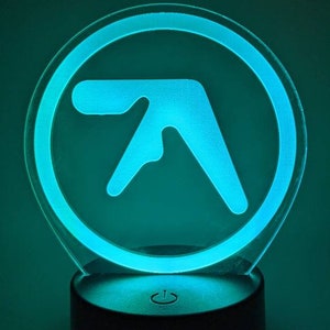 May include: A teal blue night light with a white outline of the letter "A" inside a circle. The night light has a black base with a power button.