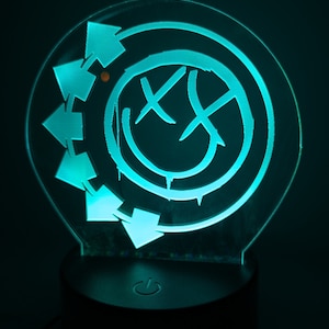 May include: A teal blue 3D printed night light with a smiley face with an X over each eye and a circle of arrows around the outside. The night light has a black base with a power button.