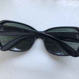May include: Black sunglasses with the Ray-Ban logo on the temples. The sunglasses have a thick, rectangular frame and dark lenses. The arms are folded, showing the interior of the glasses.