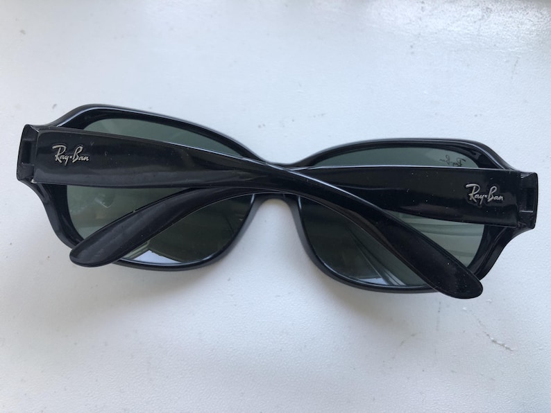 May include: Black sunglasses with the Ray-Ban logo on the temples. The sunglasses have a thick, rectangular frame and dark lenses. The arms are folded, showing the interior of the glasses.