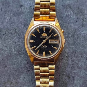 May include: A gold-tone Orient watch with a black face and a gold metal band. The watch face has a date window and the text "AAA Crystal 21 Jewels" printed on it.