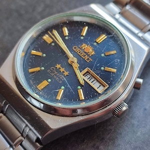 May include: A silver Orient Crystal 21 Jewels wristwatch with a blue face, gold hands, and a day of the week display that reads "FRI 1".