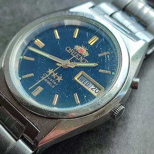 May include: A silver Orient Crystal 21 Jewels watch with a blue face, gold hands, and a date window that reads "WED 20".