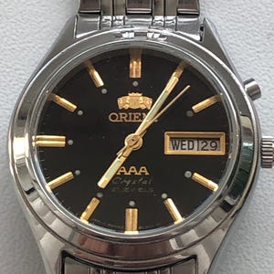 May include: A silver wristwatch with a black face and gold accents. The watch face has a day and date window, and the text "AAA Crystal 21 Jewels" is printed on the face. The watch has a silver metal band.