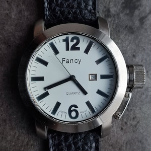 May include: A silver-toned wristwatch with a white face, black numerals, and hands. The watch face reads "Fancy" and "Quartz." The black leather band is textured. The date is displayed on the watch face.