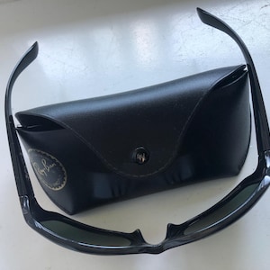 May include: Black sunglasses with a matching black case. The sunglasses have a sleek, modern design with dark lenses and a curved frame. The case is rectangular and features a button closure and a logo.