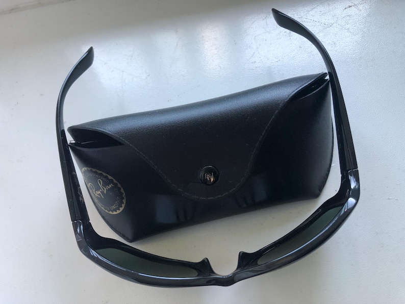 May include: Black sunglasses with a matching black case. The sunglasses have a sleek, modern design with dark lenses and a curved frame. The case is rectangular and features a button closure and a logo.