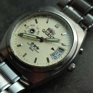 May include: A silver wristwatch with a white face and black hands. The watch has a date window and a day of the week window. The watch is water resistant to 50 meters. The watch has the words "King Orient" on the face.