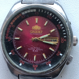 May include: A vintage Orient automatic wristwatch with a stainless steel band and case. The watch face is a deep red with gold-colored hands and markers. The bezel is black and red, with white numerals. The watch has a date display at the 3 o'clock position.