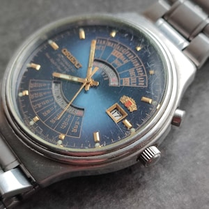 May include: A blue and silver wristwatch with a calendar dial and a gold-colored second hand. The watch face features a day of the week, date, and month display. The watch is made by the brand "Citizen".