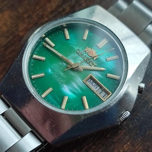May include: A silver wristwatch with a green face and a silver band. The watch face has gold accents and a date window that reads "MON 11".