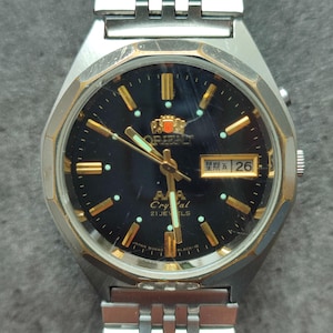 May include: A black-faced wristwatch with gold accents and a silver metal band. The watch face has a date window that reads "曜日 26" and the words "Crystal 21 Jewels" are printed below the Orient logo.