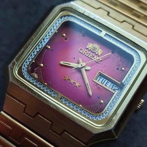 May include: A gold-toned Orient watch with a square face and a matching gold band. The watch face is a gradient of pink and purple with gold hands and markers. The day is displayed as "TUE 12".