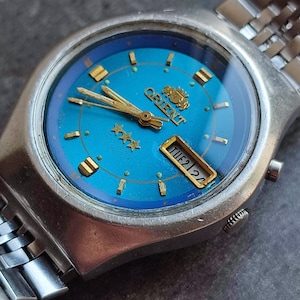 May include: A silver Orient watch with a blue face and gold accents. The watch has a day and date window and three stars on the face. The watch is on a silver metal band.