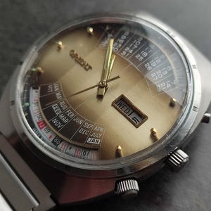 May include: A silver wristwatch with a gold face and a calendar dial. The watch face has a day of the week indicator, a date indicator, and a month indicator. The watch has a silver band.