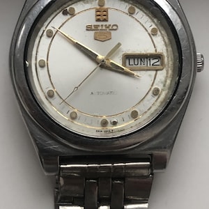 May include: A vintage Seiko 5 automatic wristwatch with a silver-tone case and band. The watch face is white with gold-tone hands and markers. The day and date are displayed in a window at the 3 o'clock position. The watch is showing the date as LUN 12.