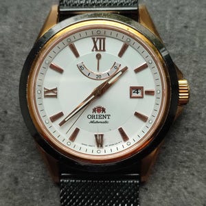 May include: A rose gold and black Orient automatic wristwatch with a white face and black mesh band. The watch face has roman numeral hour markers and a date window at the 4 o'clock position.