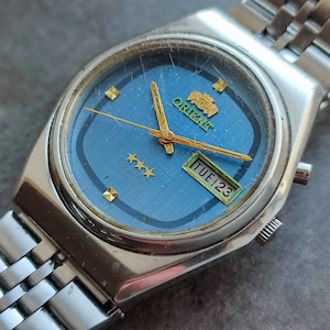 May include: A silver Orient watch with a blue face, gold hands, and a day of the week display. The watch face has three gold stars and the text "TUE 23".