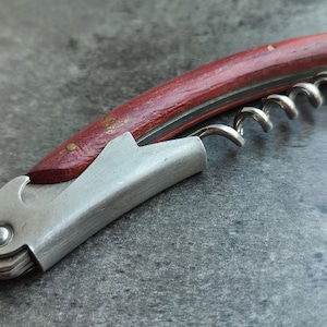 Pode incluir: Um saca-rolhas com cabo de madeira vermelha e corpo de metal prateado, com um saca-rolhas em espiral.