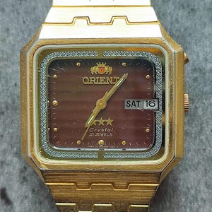 May include: A gold-toned Orient watch with a brown face and a gold metal band. The watch face has a date window that reads "SAT 16" and the text "Crystal 21 Jewels".