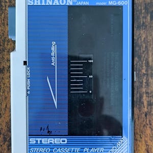 May include: A Shinaon Japan model MG-600 stereo cassette player with a blue and black faceplate. The device features a clear window displaying a cassette tape labeled The Bodyguard, with printed text including Anti-Rolling and Push Lock.